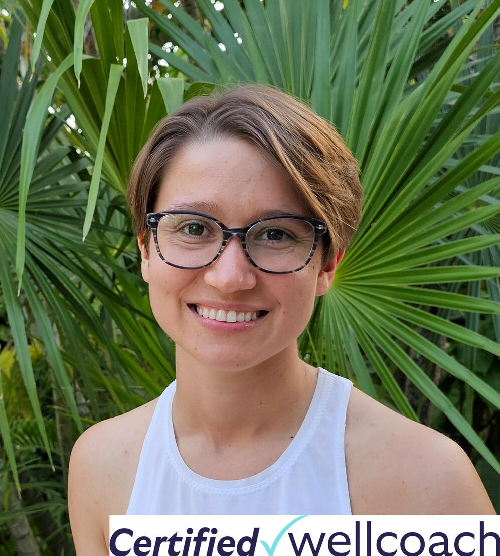 Image of Krysta Liz, short brown hair parted, smiling, wearing glasses, a white tank top, in front of a palmetto bush