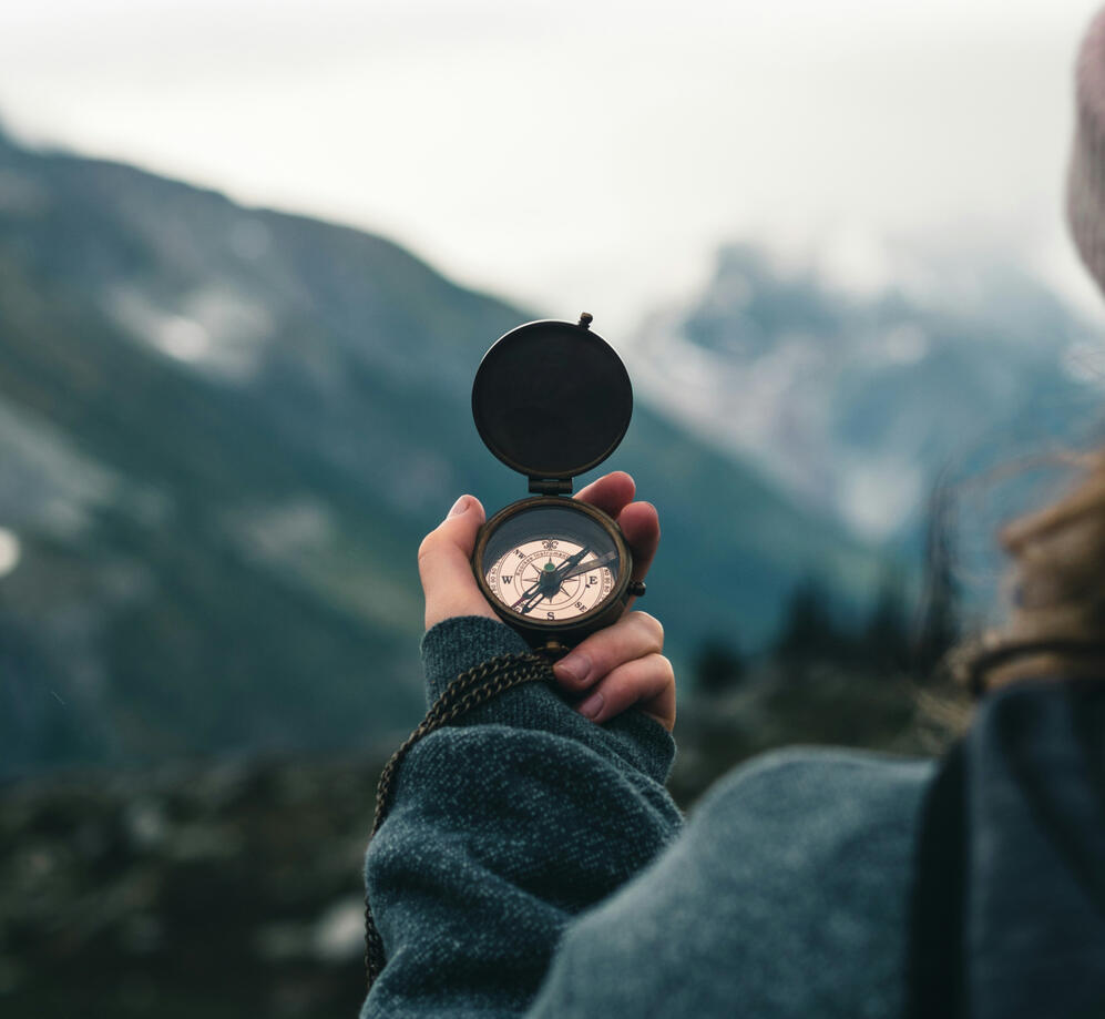 Image of a compass held in the left hand of a person with a blurry mountain forest background