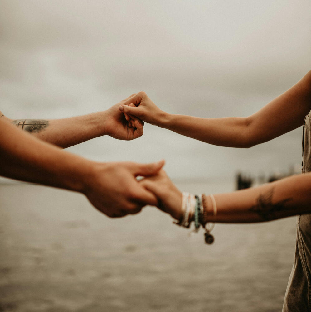 Image of people's arms holding hands in a circle as if dancing or praying, blurry overcast beach background